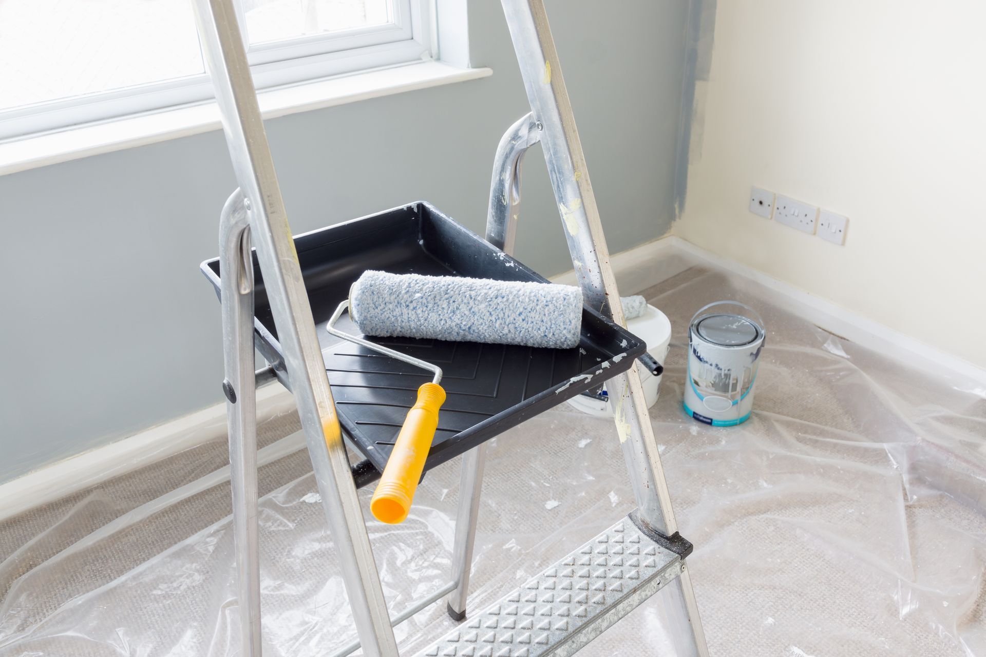 A paint roller and tray sit on a step ladder in a room being painted, with a paint can on the floor.