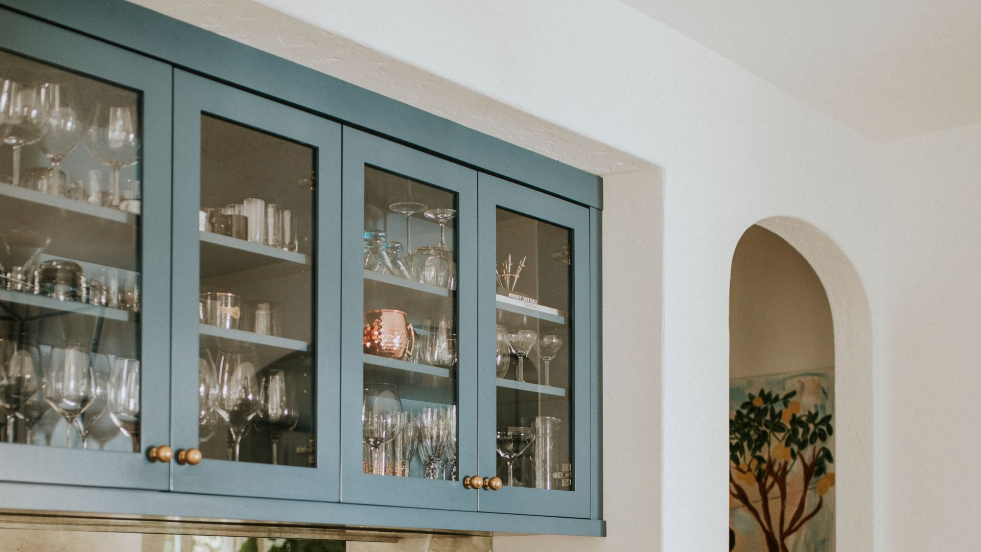 Blue-painted upper cabinets with glass doors, displaying glassware and dishes, next to a white arched doorway.