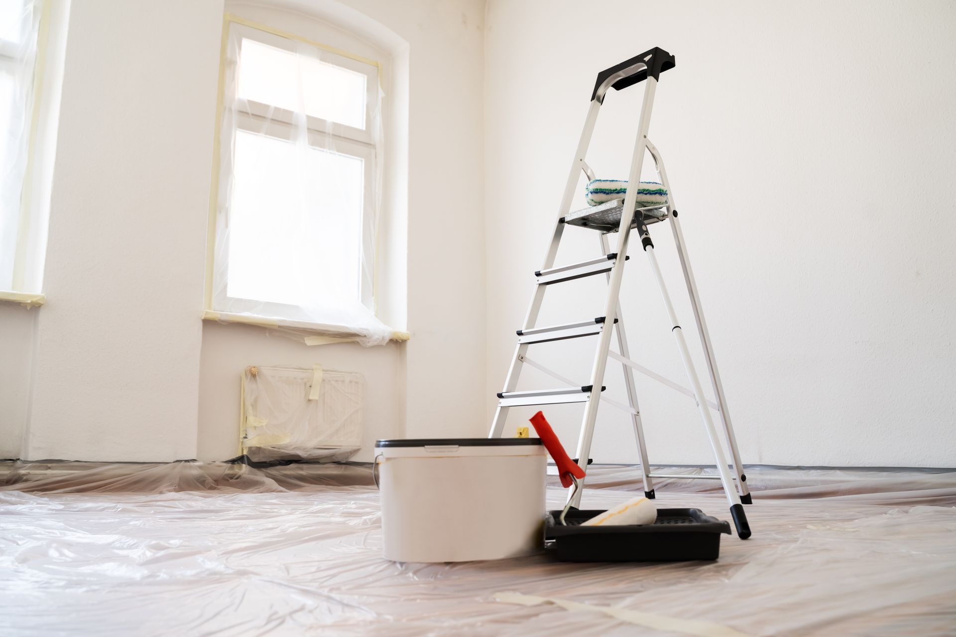 Room being painted, with ladder, paint bucket, and roller. White walls, plastic floor covering.