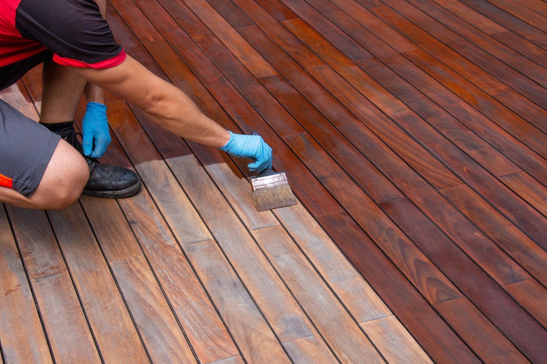 Person applying wood stain to a wooden deck with a brush; rich, reddish-brown stain contrasts with unfinished wood.