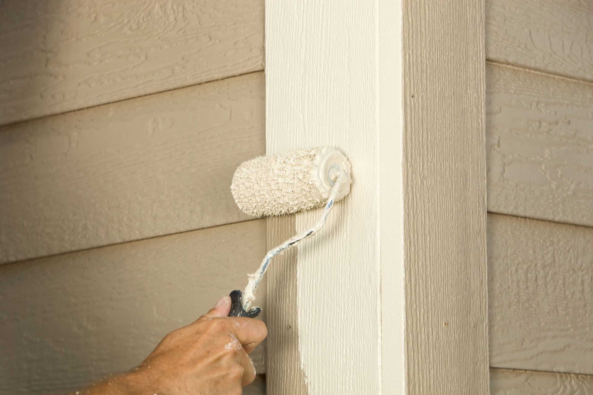 A hand using a paint roller on a light-colored pillar; siding in the background.