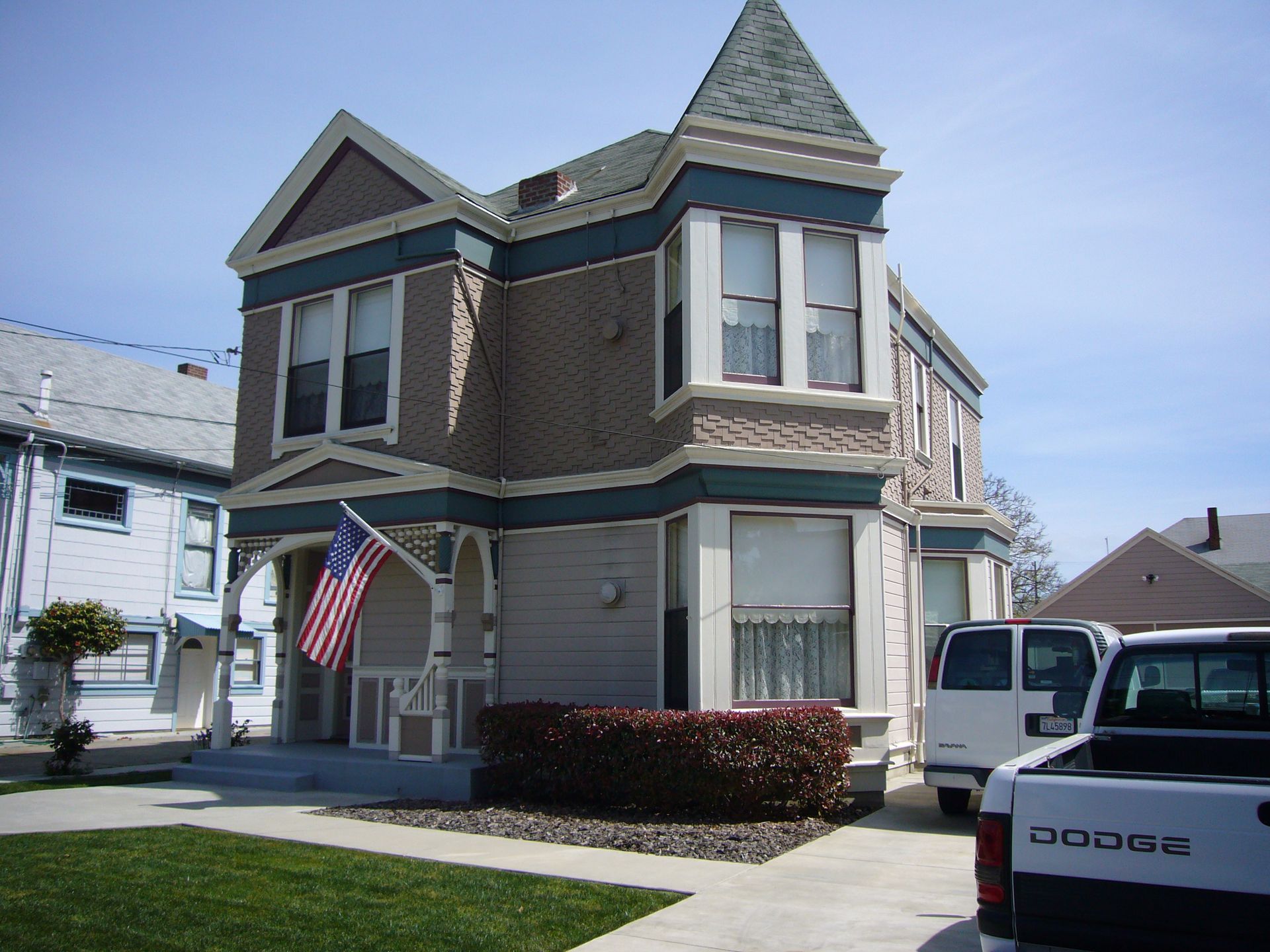 Victorian house with American flag, beige and blue accents, parked white van and truck.