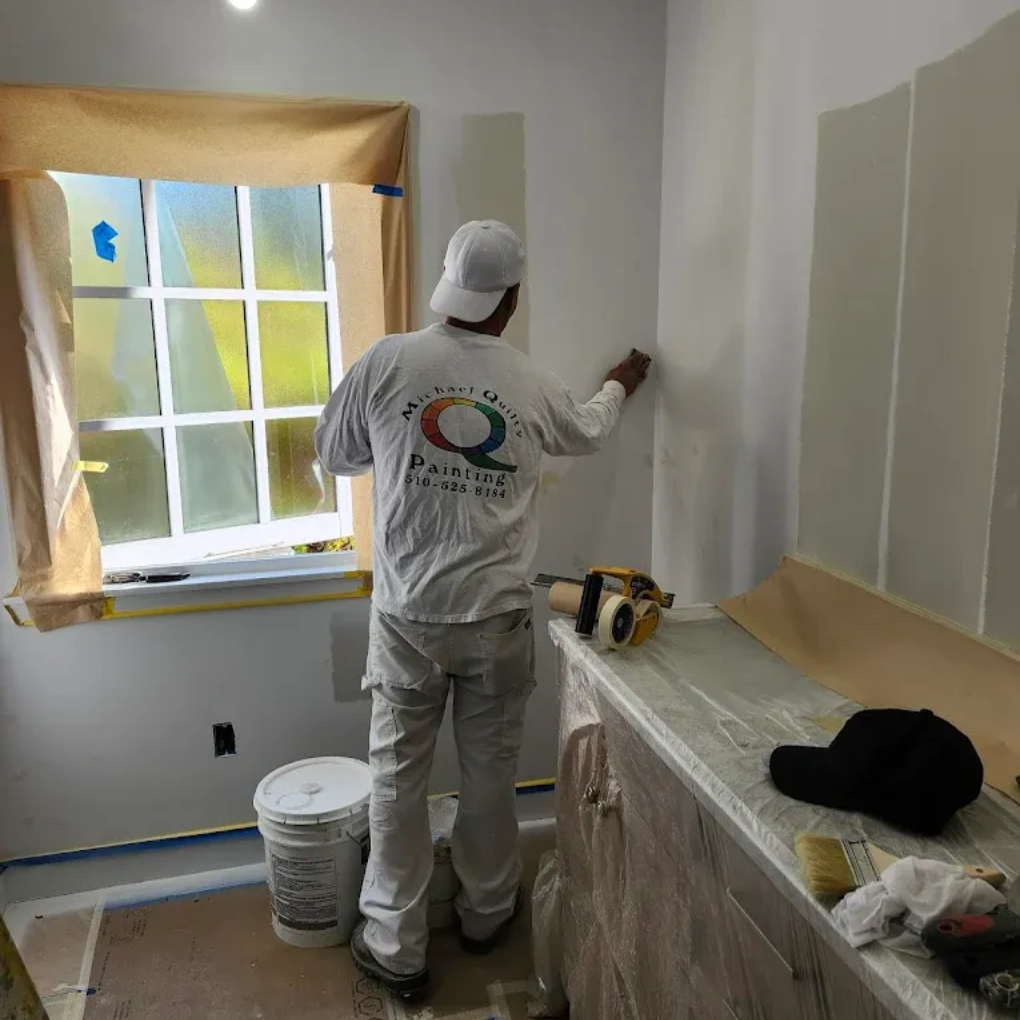 Painter applying paint to a wall. Room setting, window with covering, tools on counter.