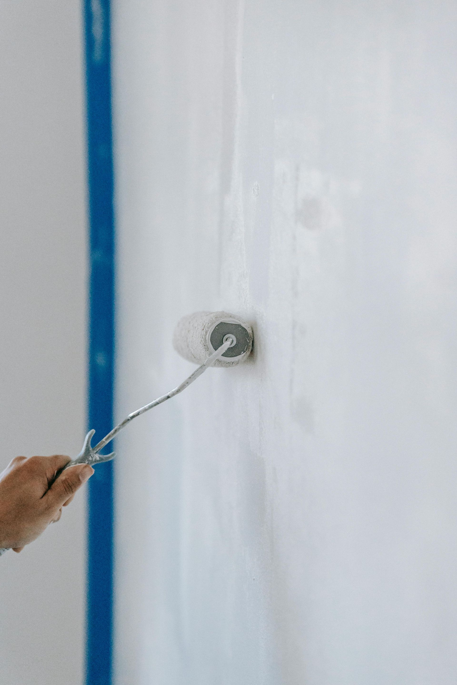 A person painting a white wall with a roller. Blue tape marks the edge.