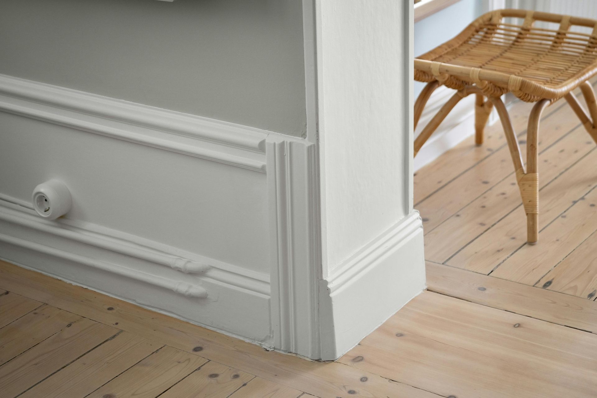 White wall molding and baseboard corner with wooden floor and woven stool.