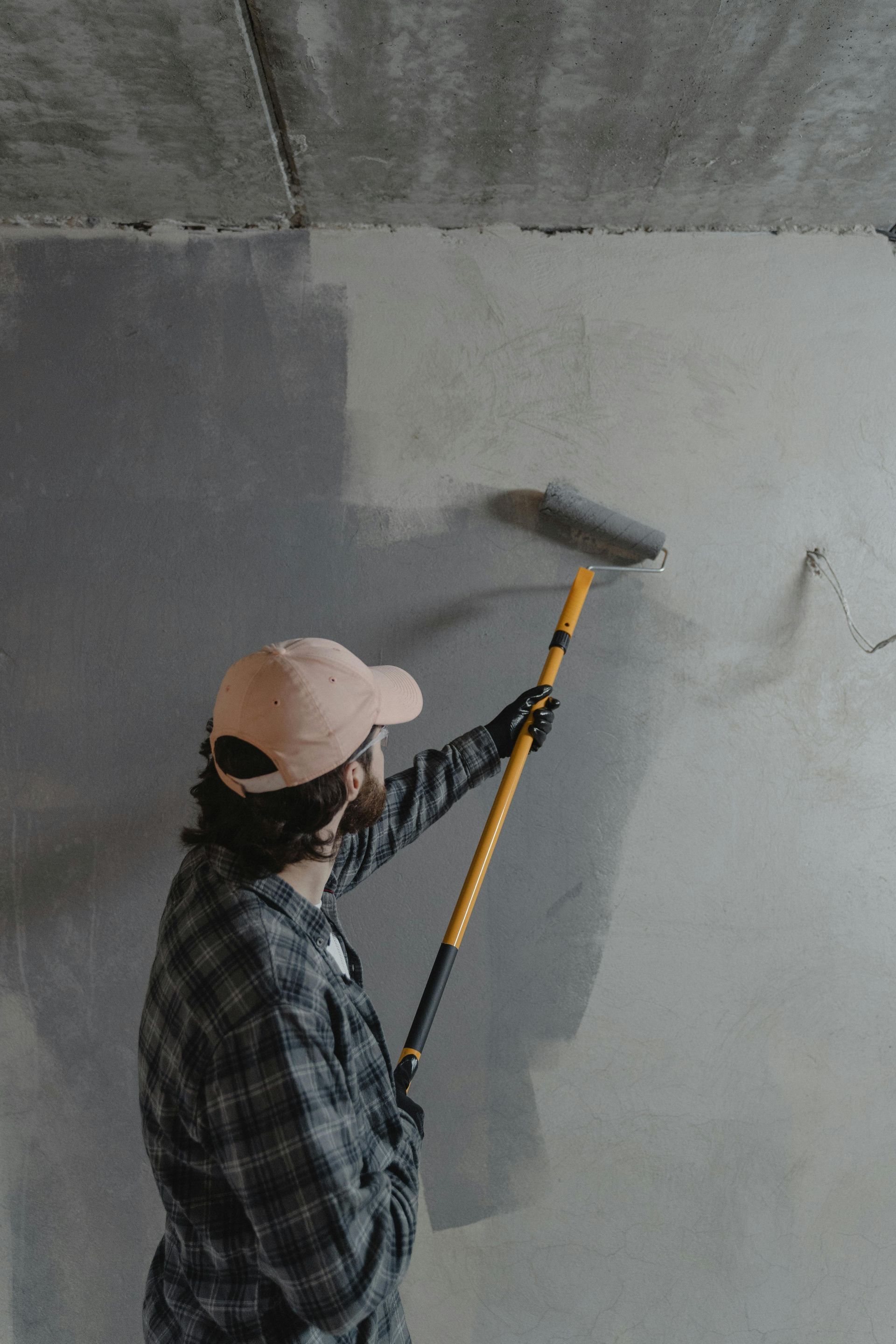 Person painting a concrete wall gray with a paint roller on a pole.