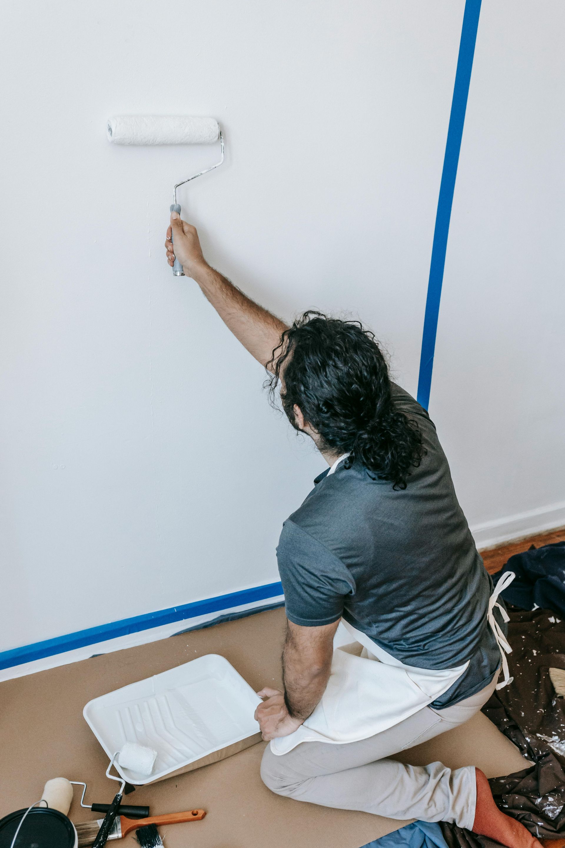 Person paints a white wall with a roller. Blue tape marks the edge. Paint tray and tools are nearby.