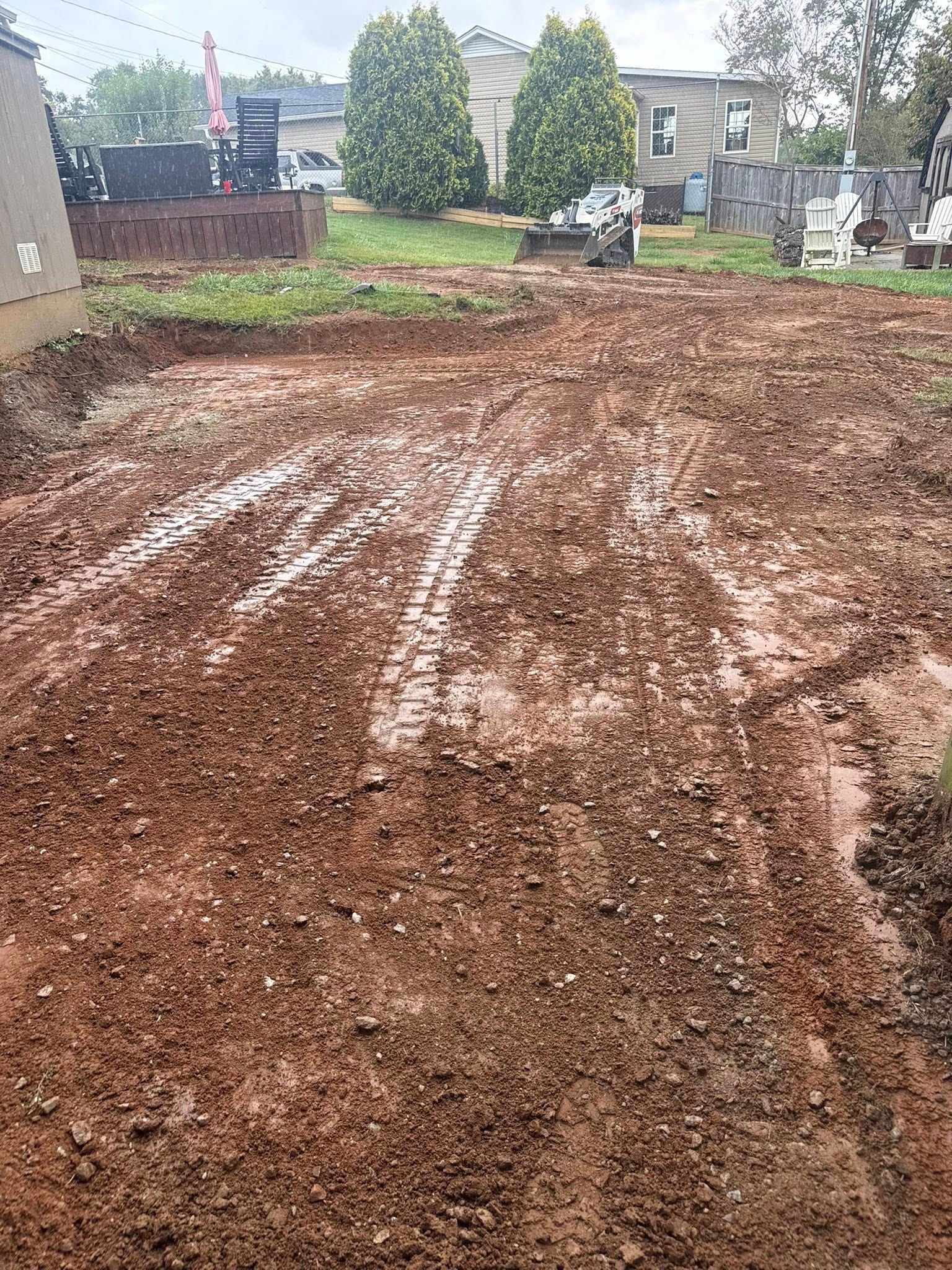 Muddy, cleared construction site with tire tracks; trees and buildings in the background.