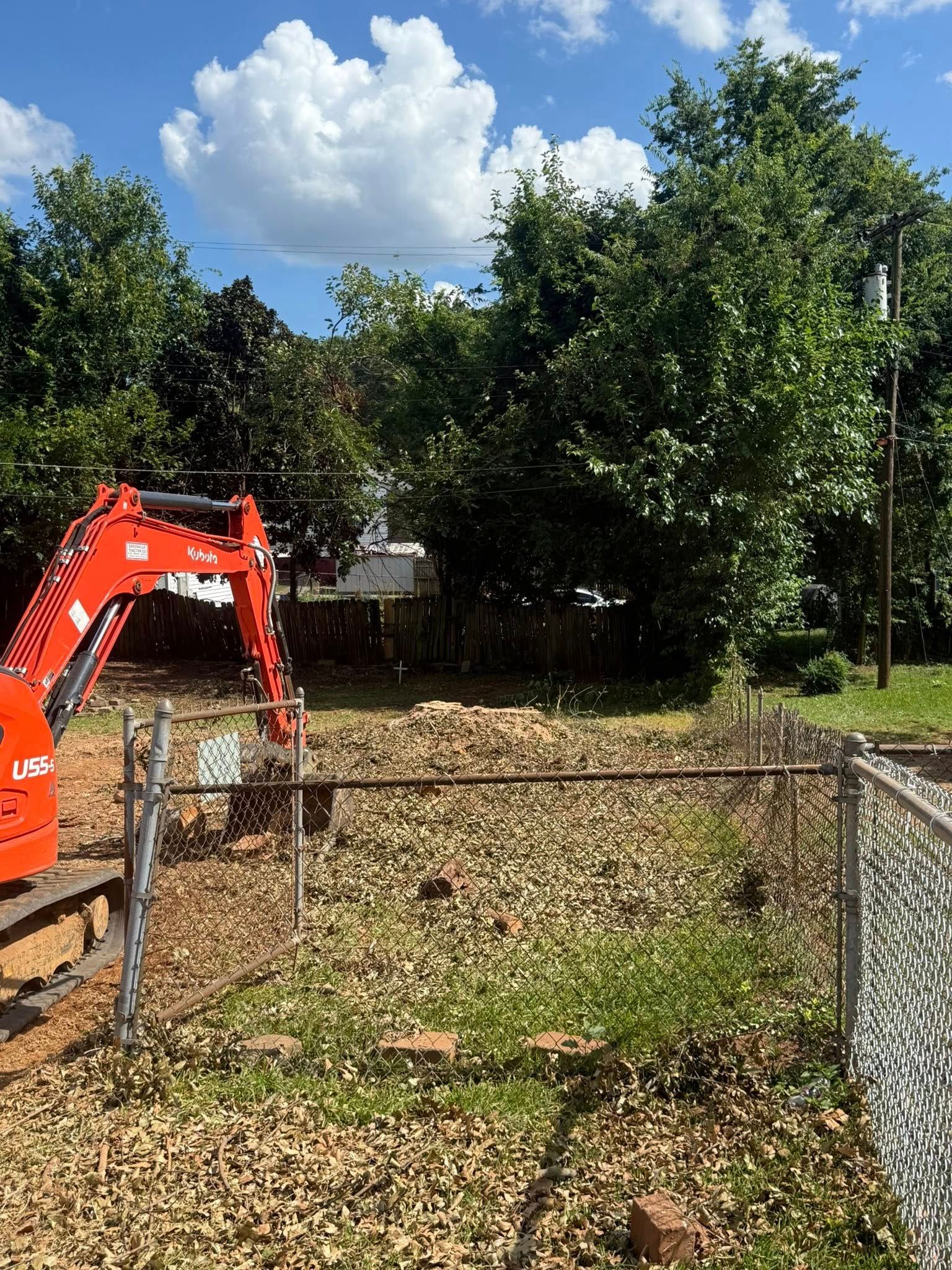A small excavator clearing debris in a fenced-in yard on a sunny day.