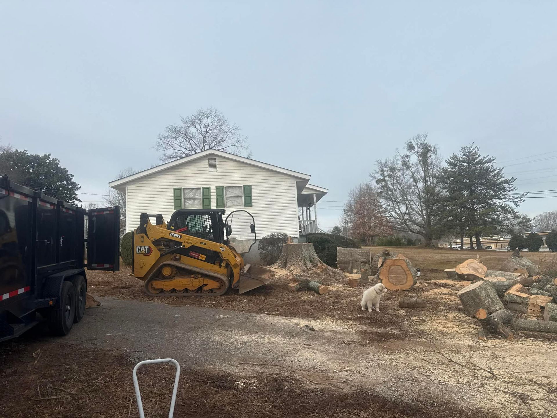 Yellow skid steer clearing debris next to a white house with green shutters and a trailer on a gravel driveway.