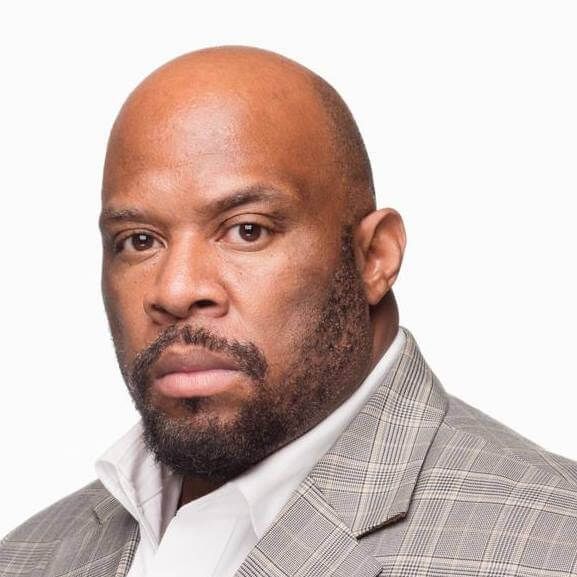 Bald man in a grey suit and open white collared shirt looking directly at the camera.