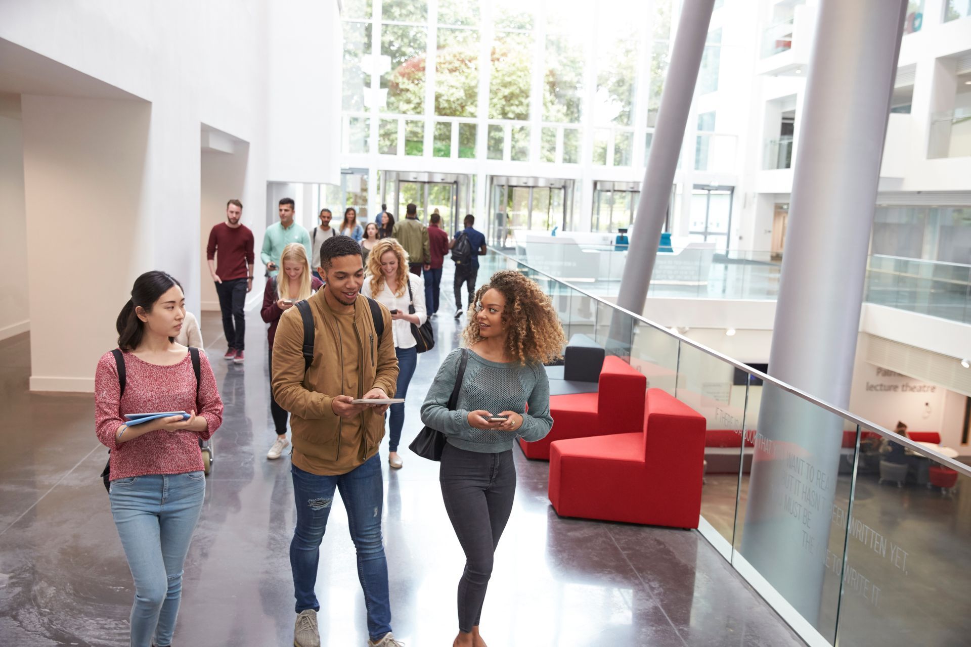 Students walking in a modern building's hallway; red seating, natural light, and tall windows.