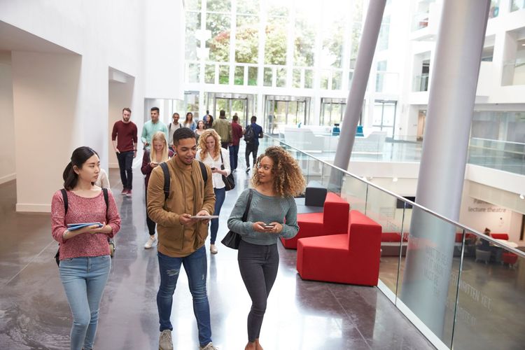 Students walking in a modern building's hallway; red seating, natural light, and tall windows.