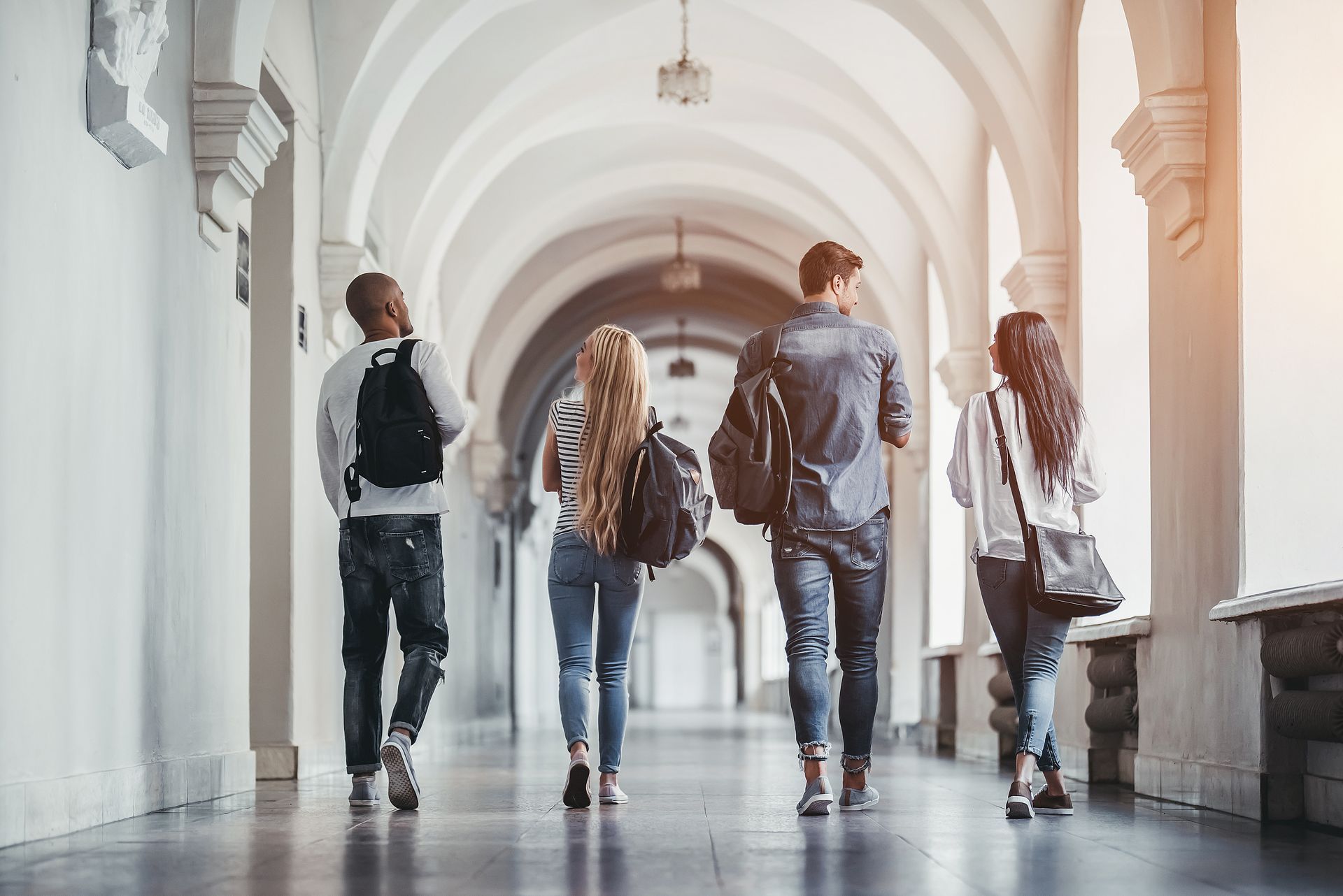 Four students walking down a sunlit arched hallway, carrying bags. Four students walking down a sunlit arched hallway, carrying bags.