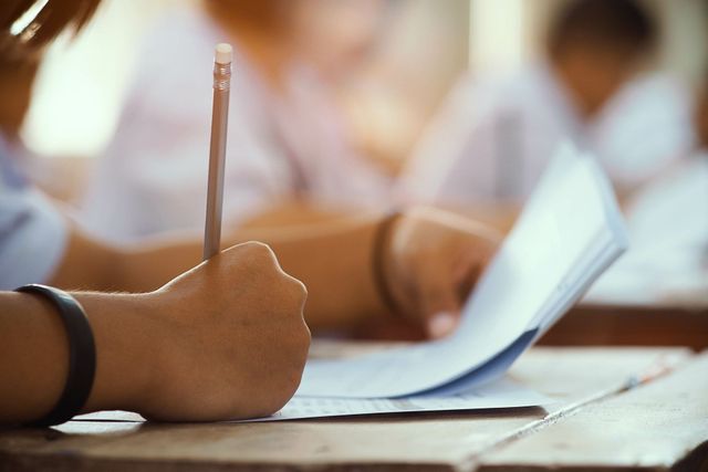 Hand holding a pencil, writing on a paper. In a classroom setting, blurred background of students. Hand holding a pencil, writing on a paper. In a classroom setting, blurred background of students.
