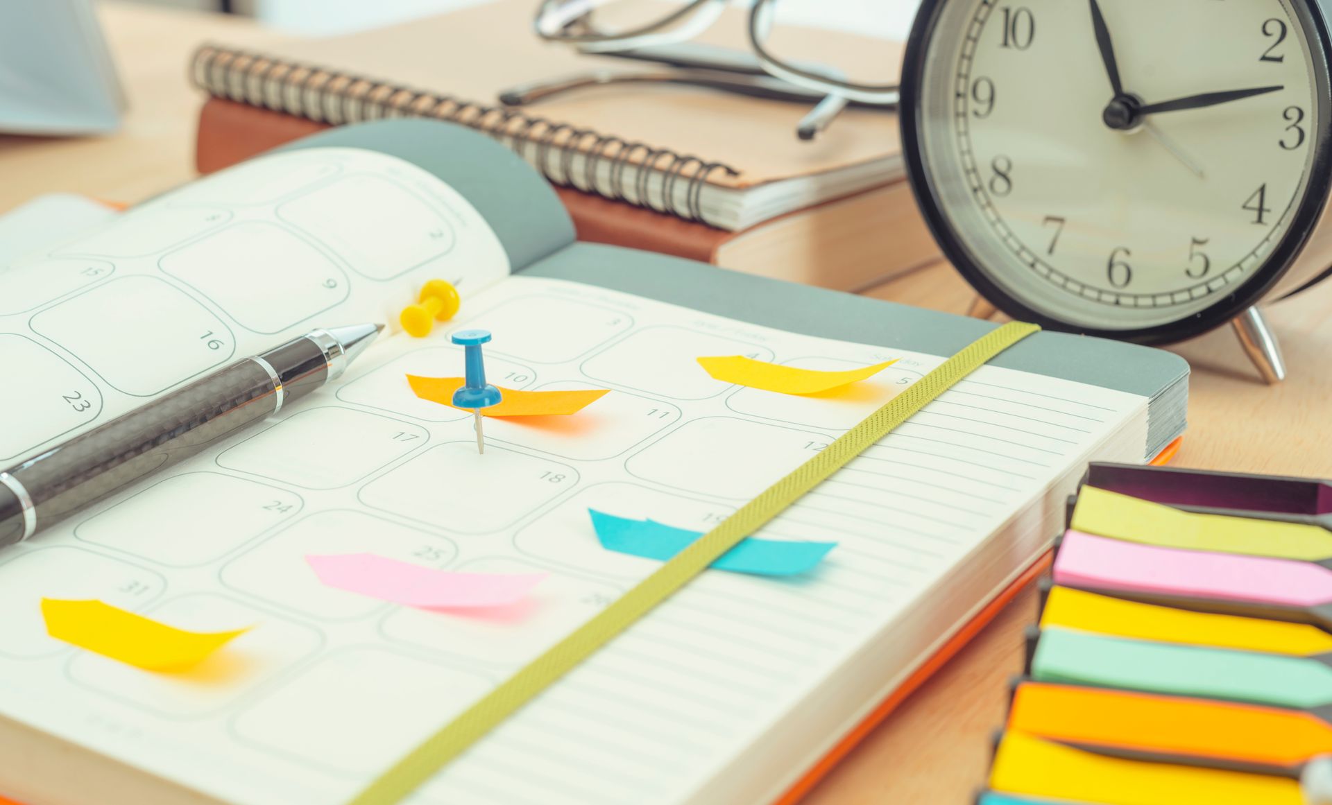 Open planner with colorful sticky notes, a pen, clock, and other office supplies on a wooden desk. Open planner with colorful sticky notes, a pen, clock, and other office supplies on a wooden desk.
