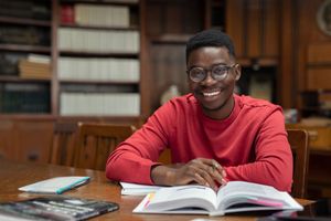 Man wearing glasses smiles at the camera, sitting at a desk with an open book in a library.