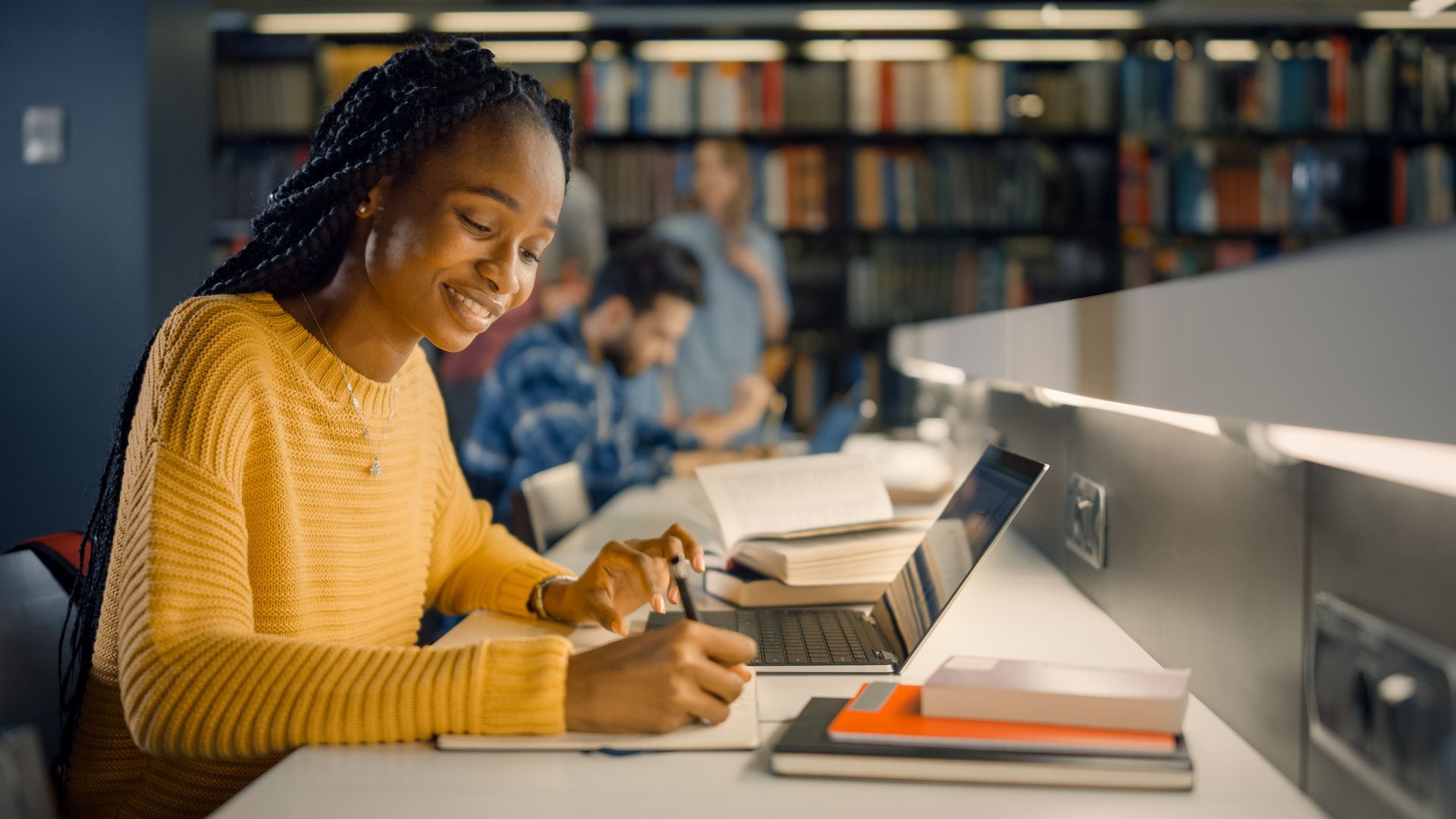 Woman in yellow sweater studies at a desk in a library, writing in a notebook, laptop open.