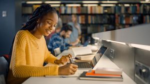 Woman in yellow sweater studies at a desk in a library, writing in a notebook, laptop open.