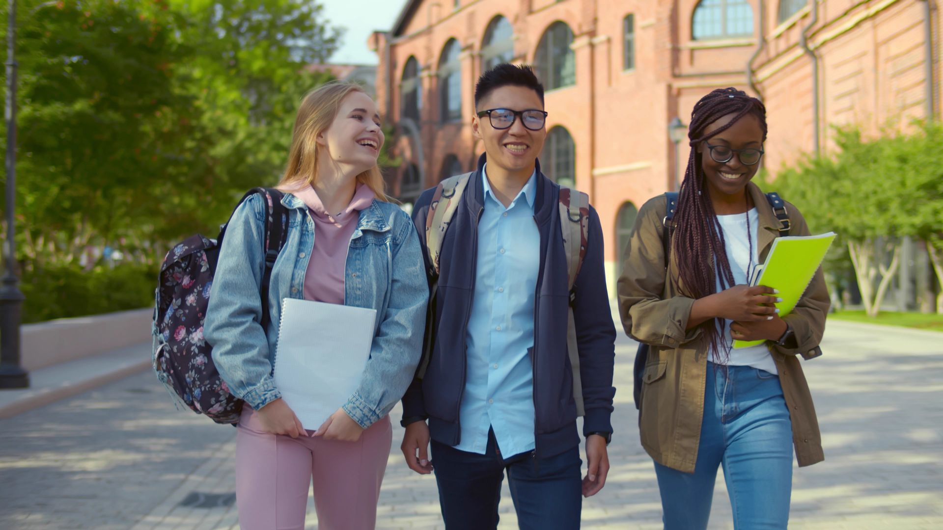 Three students walking and smiling outside a brick building, holding books and backpacks. Three students walking and smiling outside a brick building, holding books and backpacks.