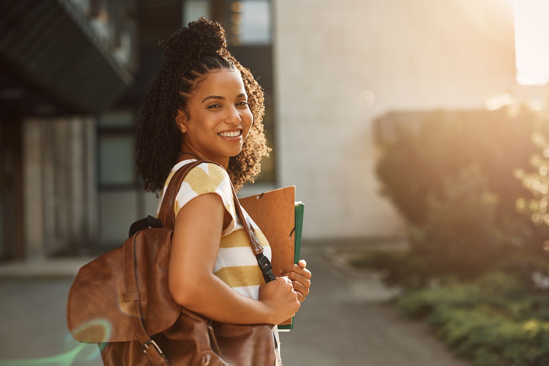 Woman with backpack and books smiles in front of a building in sunlight. Woman with backpack and books smiles in front of a building in sunlight.