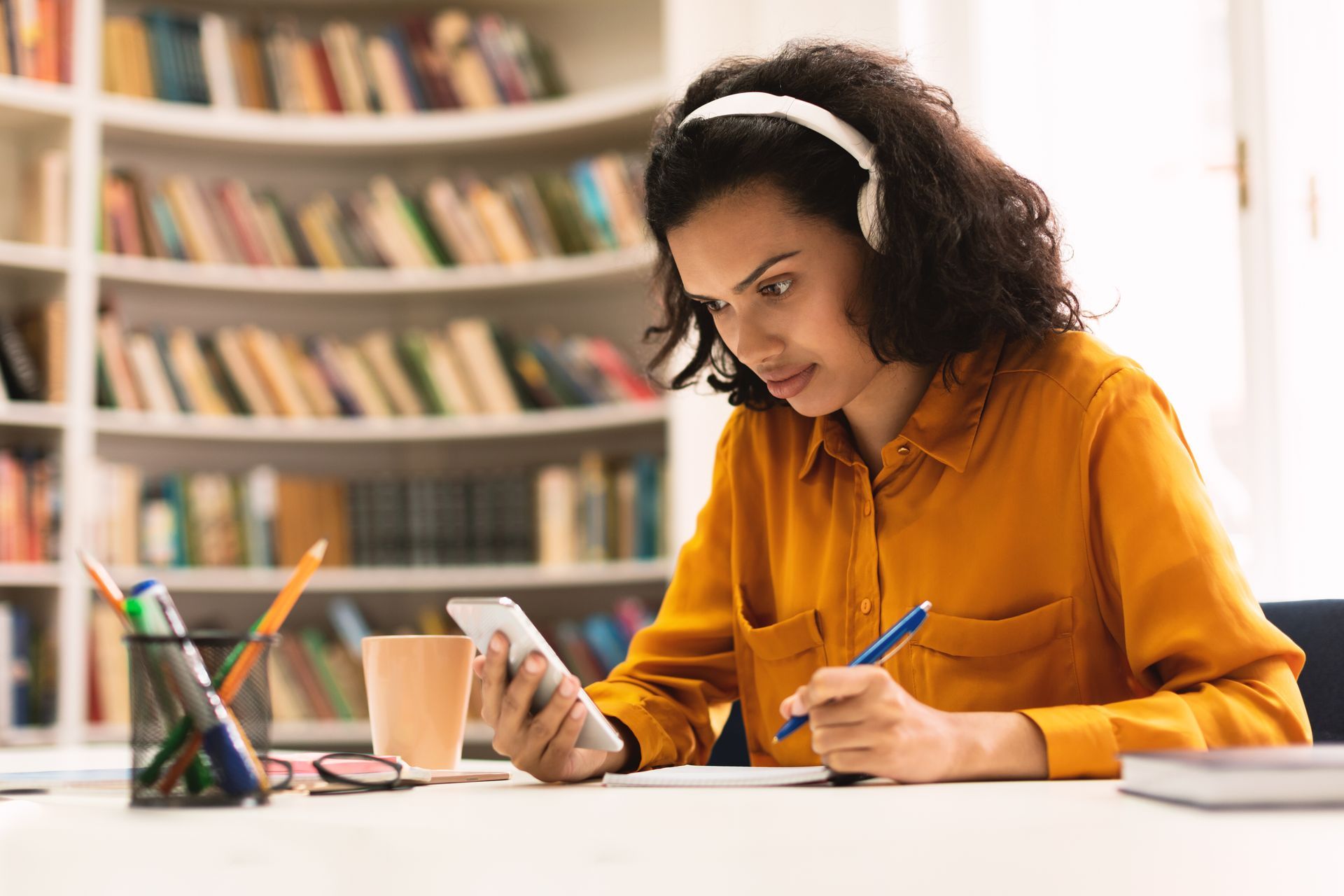 Woman wearing headphones, using a phone and taking notes at a desk in front of a bookshelf. Woman wearing headphones, using a phone and taking notes at a desk in front of a bookshelf.