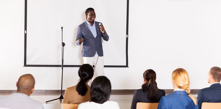 Man giving a presentation, holding a microphone, in front of an audience and a projection screen.