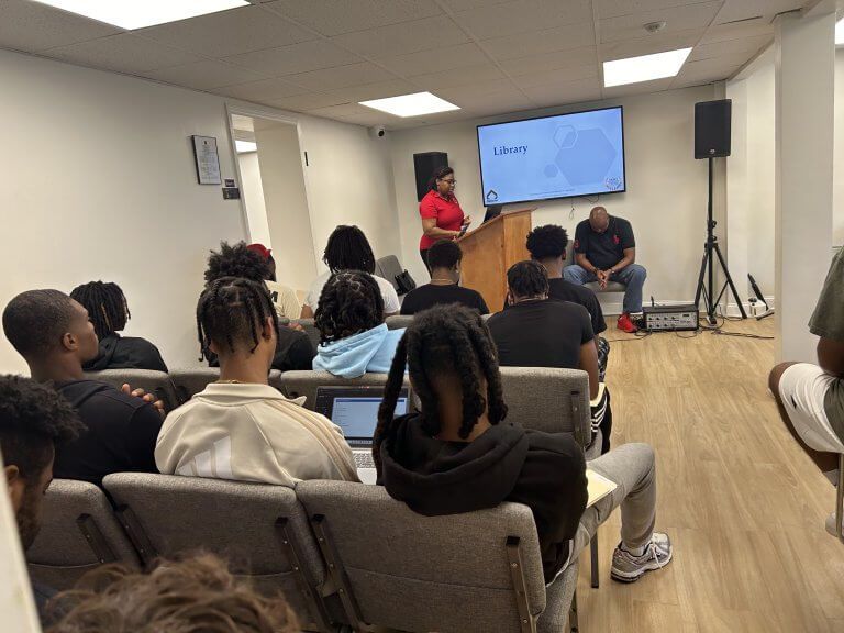 A presentation in a room. A person speaks from a podium while others listen. A screen displays text.