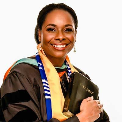 Woman in academic regalia, holding a Bible, smiling. She is wearing a yellow and blue stole.