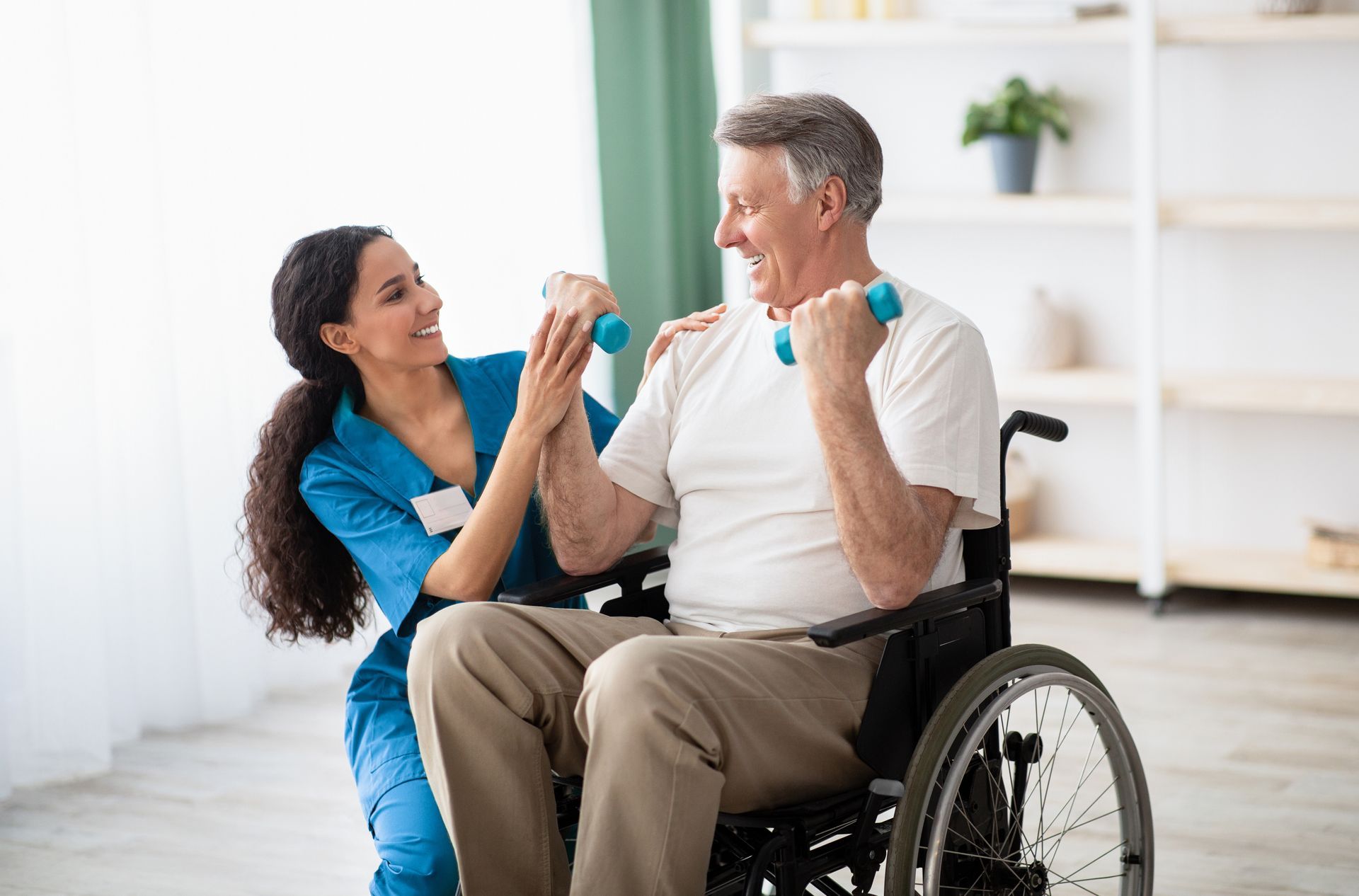 A nurse is helping an elderly man in a wheelchair lift dumbbells.