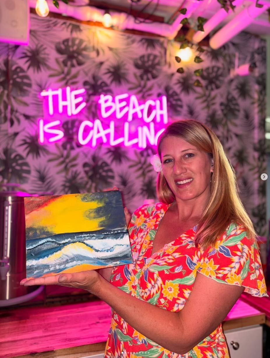A woman is holding a painting in front of a neon sign that says `` the beach is calling ''.