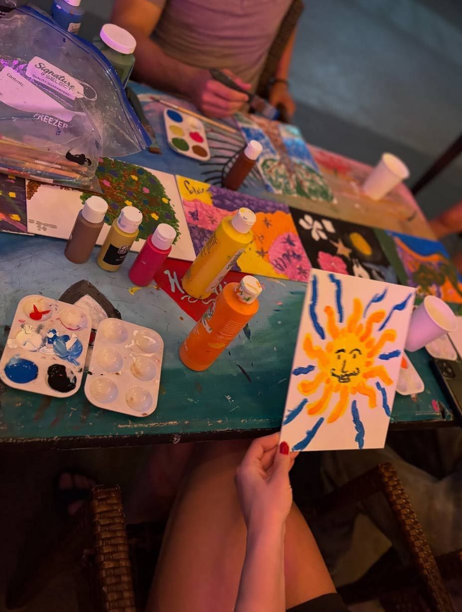 Person holding a painting of a smiling sun, surrounded by painting supplies and other artwork on a table.