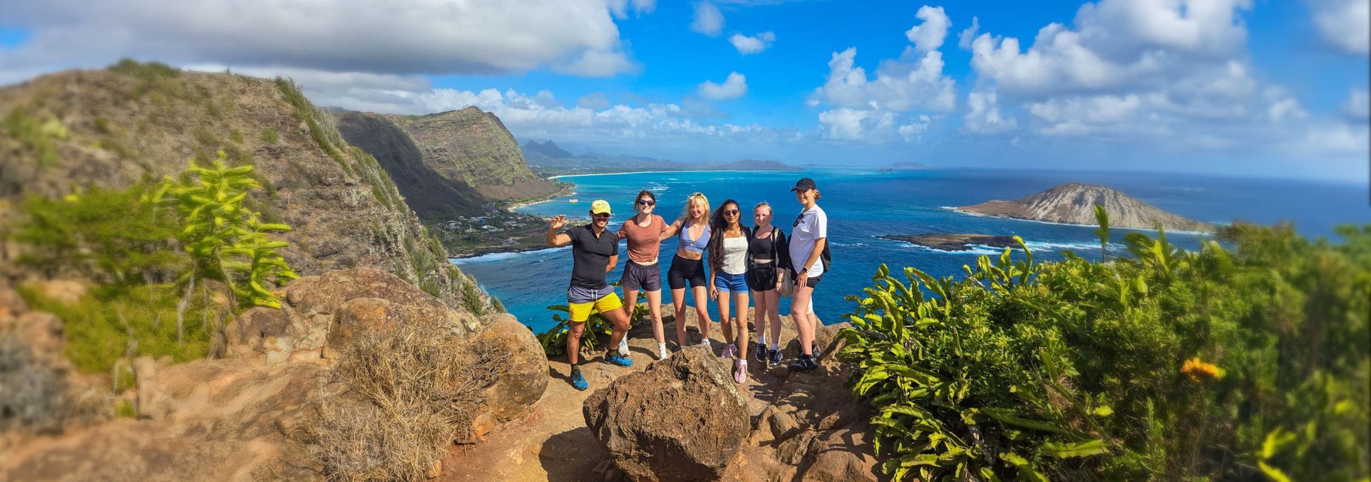A group of people poses on a cliff overlooking the ocean on a sunny day.