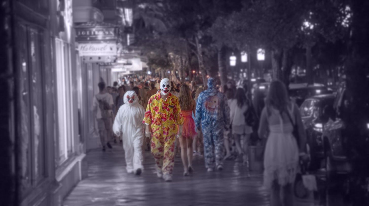 Clowns in costume walk down a busy nighttime street lined with shops and people.