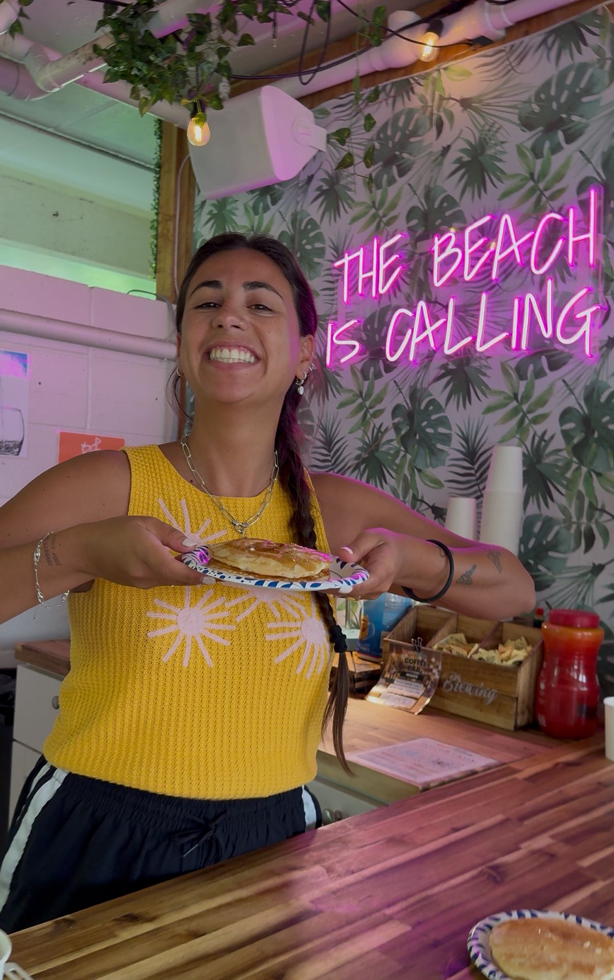 A woman is holding a plate of food in front of a neon sign that says `` the beach is calling ''.