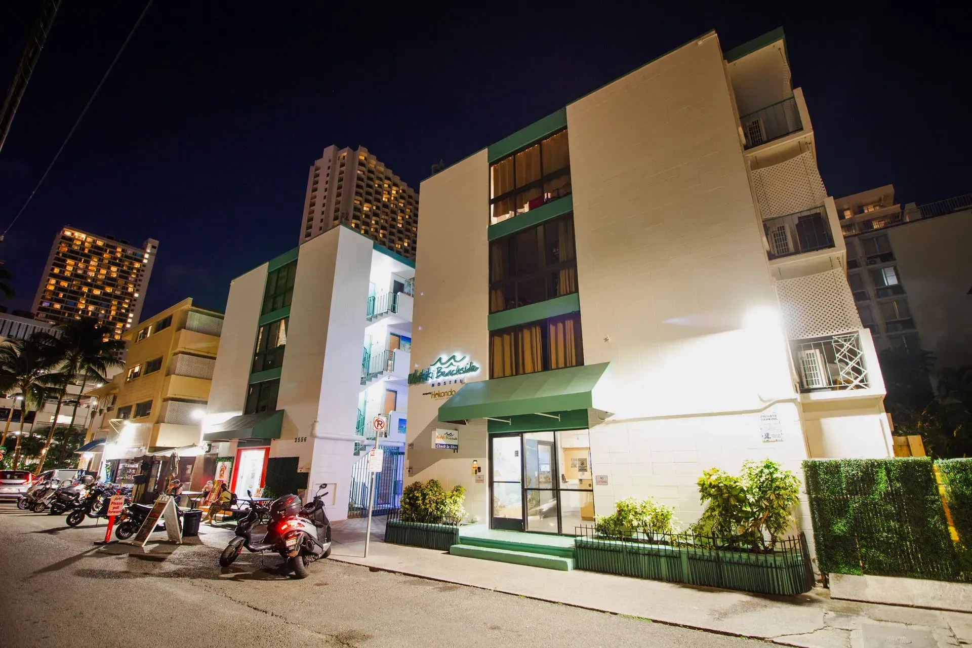 Hotel exterior at night; white and green facade, street scene with parked scooters.
