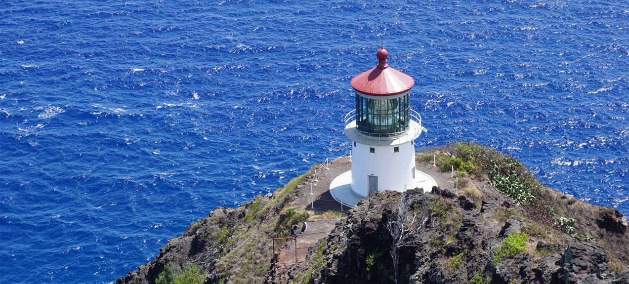 A white lighthouse with a red roof sits atop a rocky hill, surrounded by blue ocean.