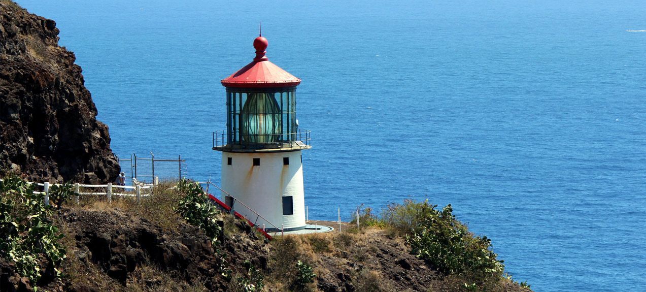 White lighthouse with a red roof sits on a rocky cliff overlooking the blue ocean.