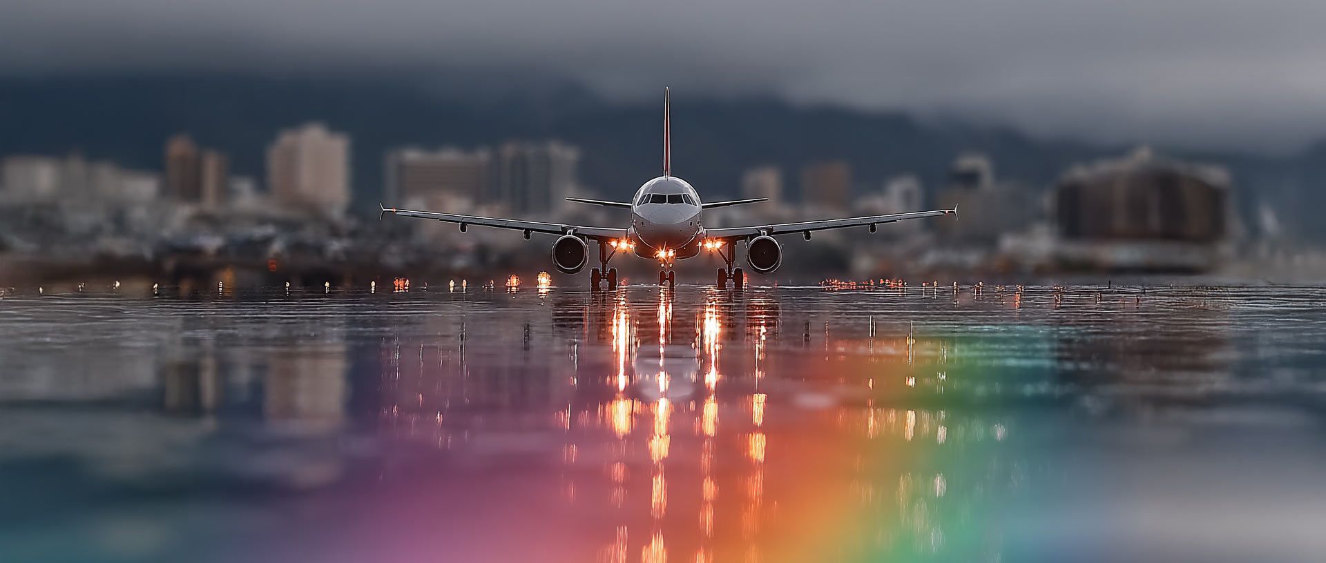 A commercial airplane taxis on a wet runway, with city buildings blurred in the background and rainbow light reflections.