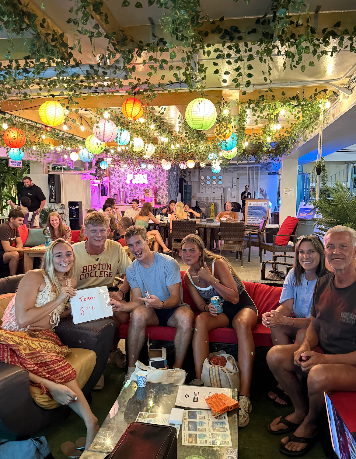Group of people smiling at a decorated outdoor cafe with colorful lanterns. Several people are on a red couch holding a sign.