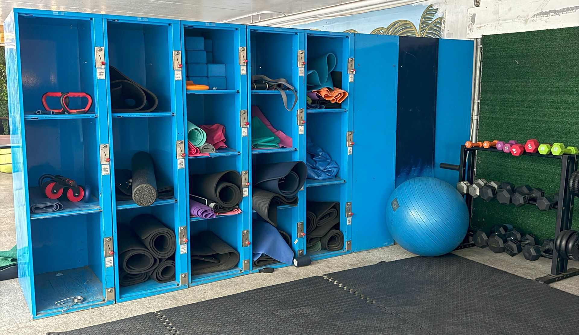 Blue storage lockers filled with exercise equipment, yoga mats, and a stability ball; next to dumbbells and weights.
