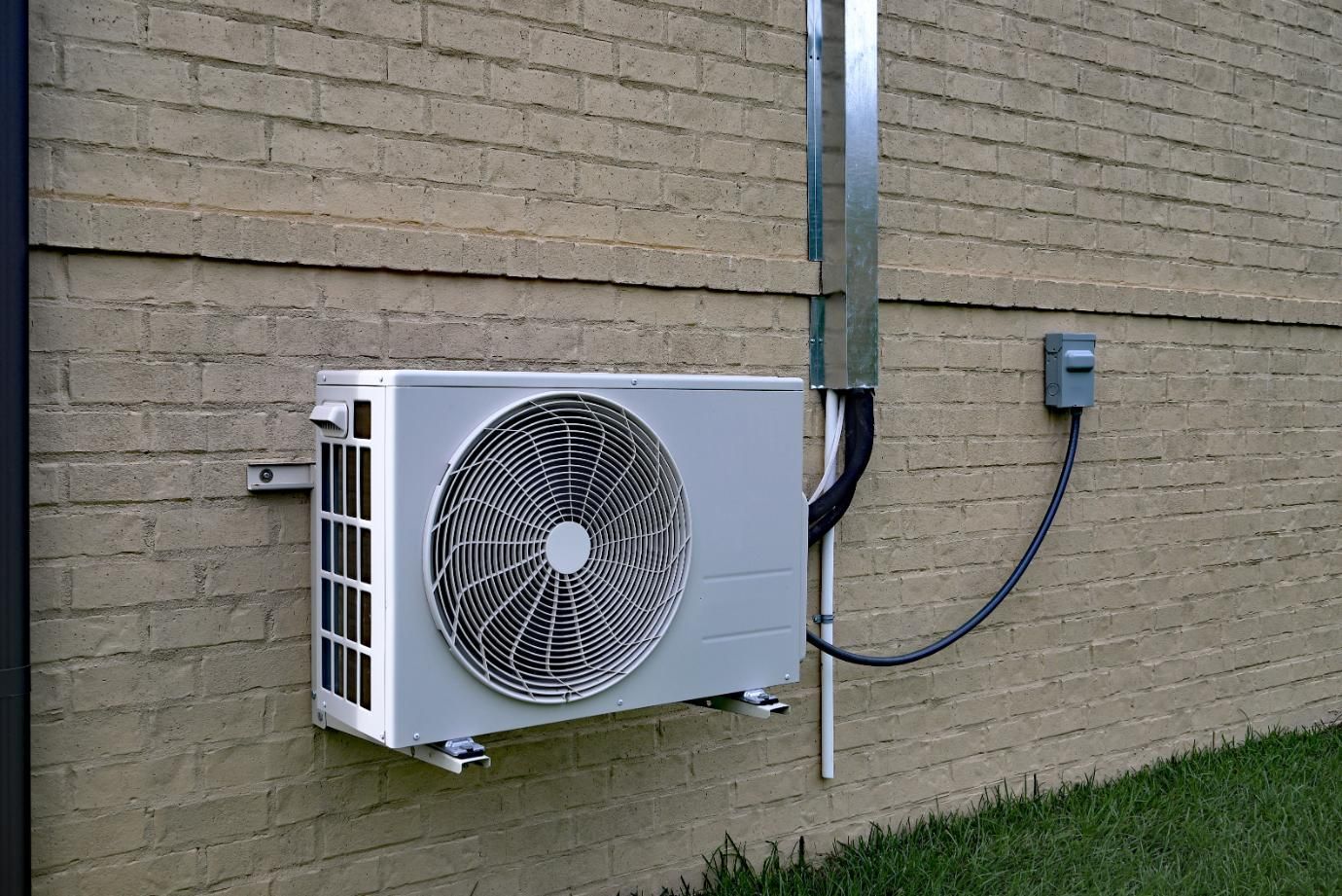 Air conditioning unit mounted on a beige brick wall with electrical conduit and outlet.