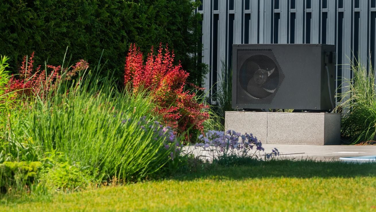 Heat pump unit on a concrete base in a lush garden, surrounded by plants and foliage.