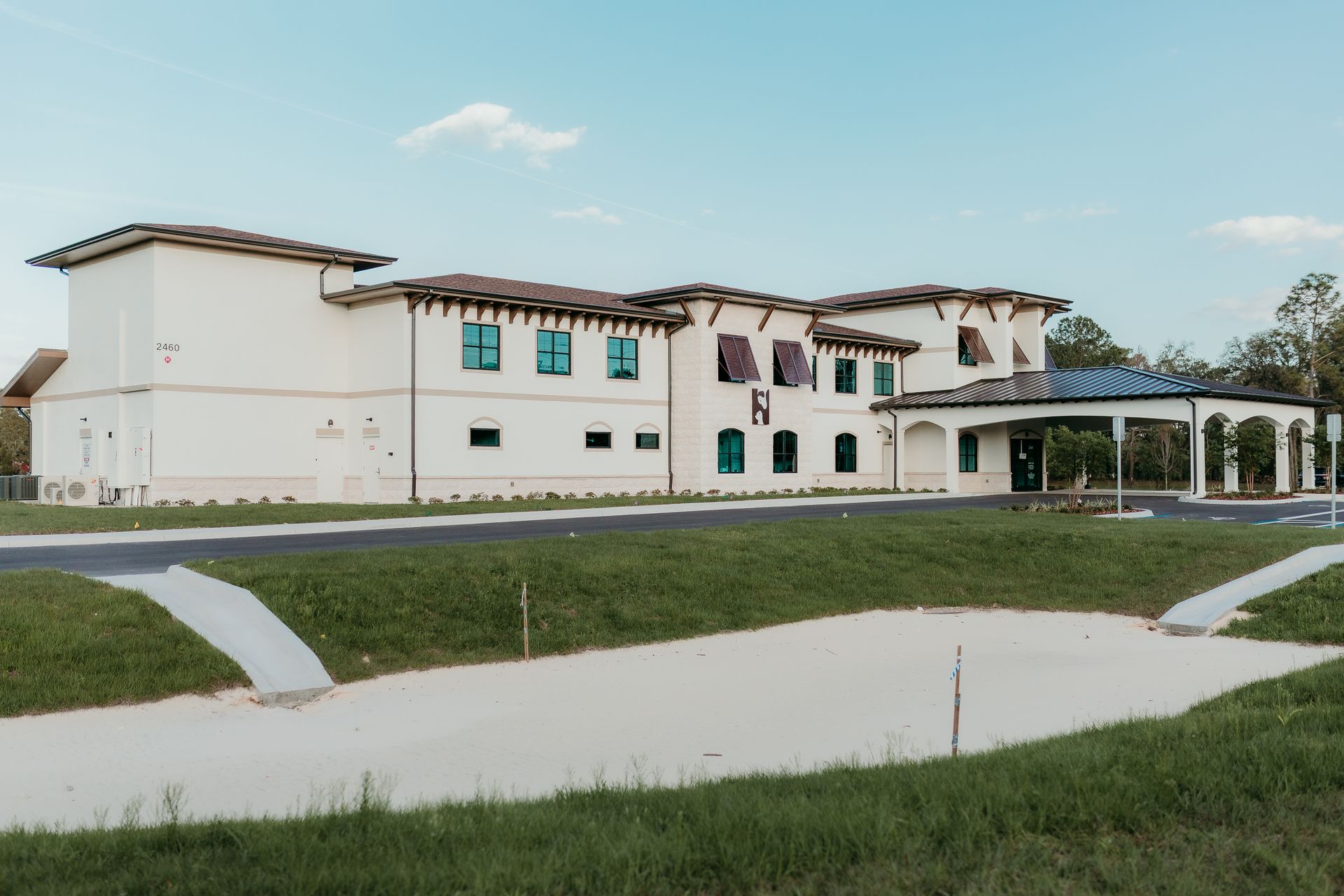 Cream-colored building with dark brown roof and green-framed windows, surrounded by green grass and a paved pathway.