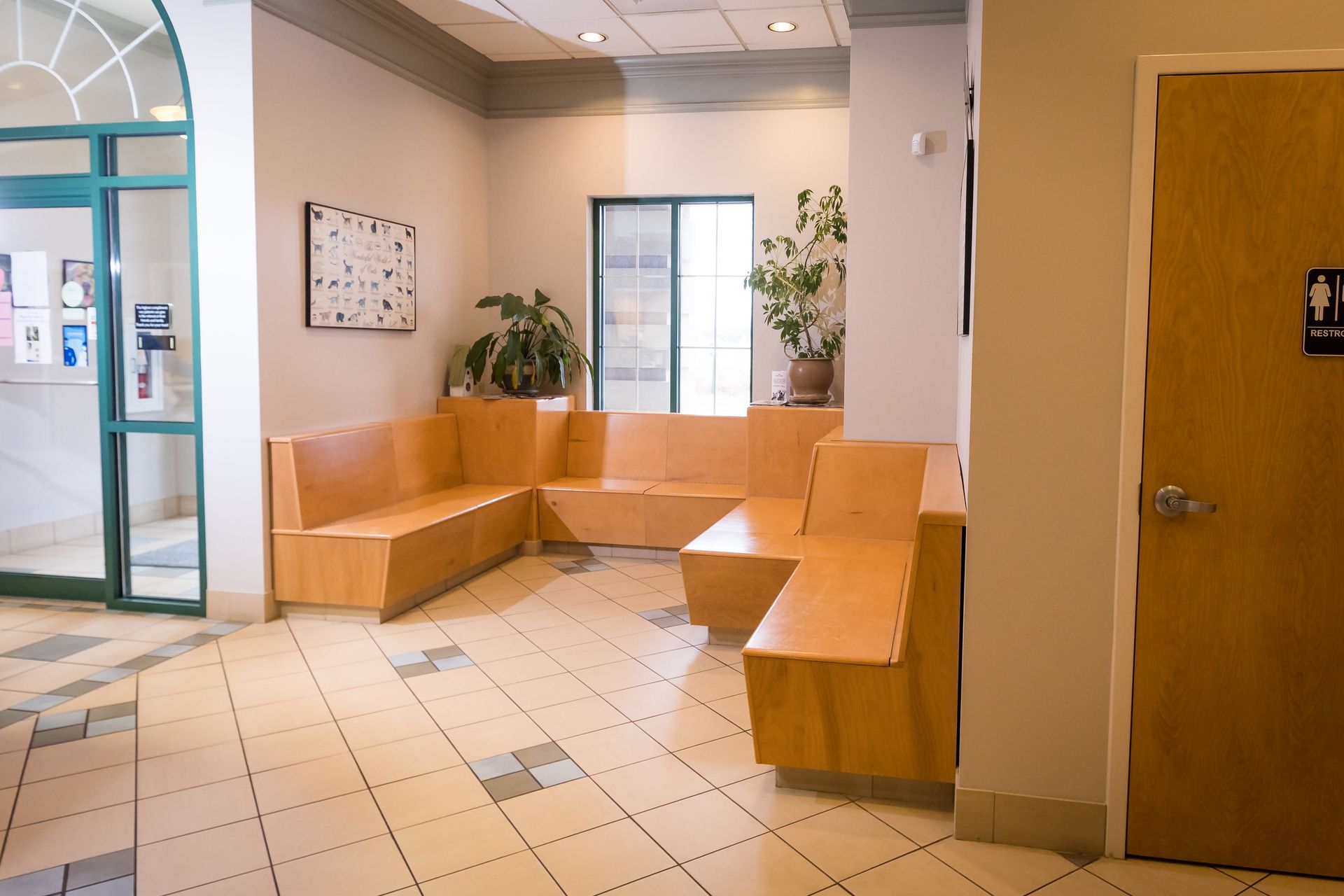 L-shaped wooden bench seating in a waiting room with plants and a restroom door.
