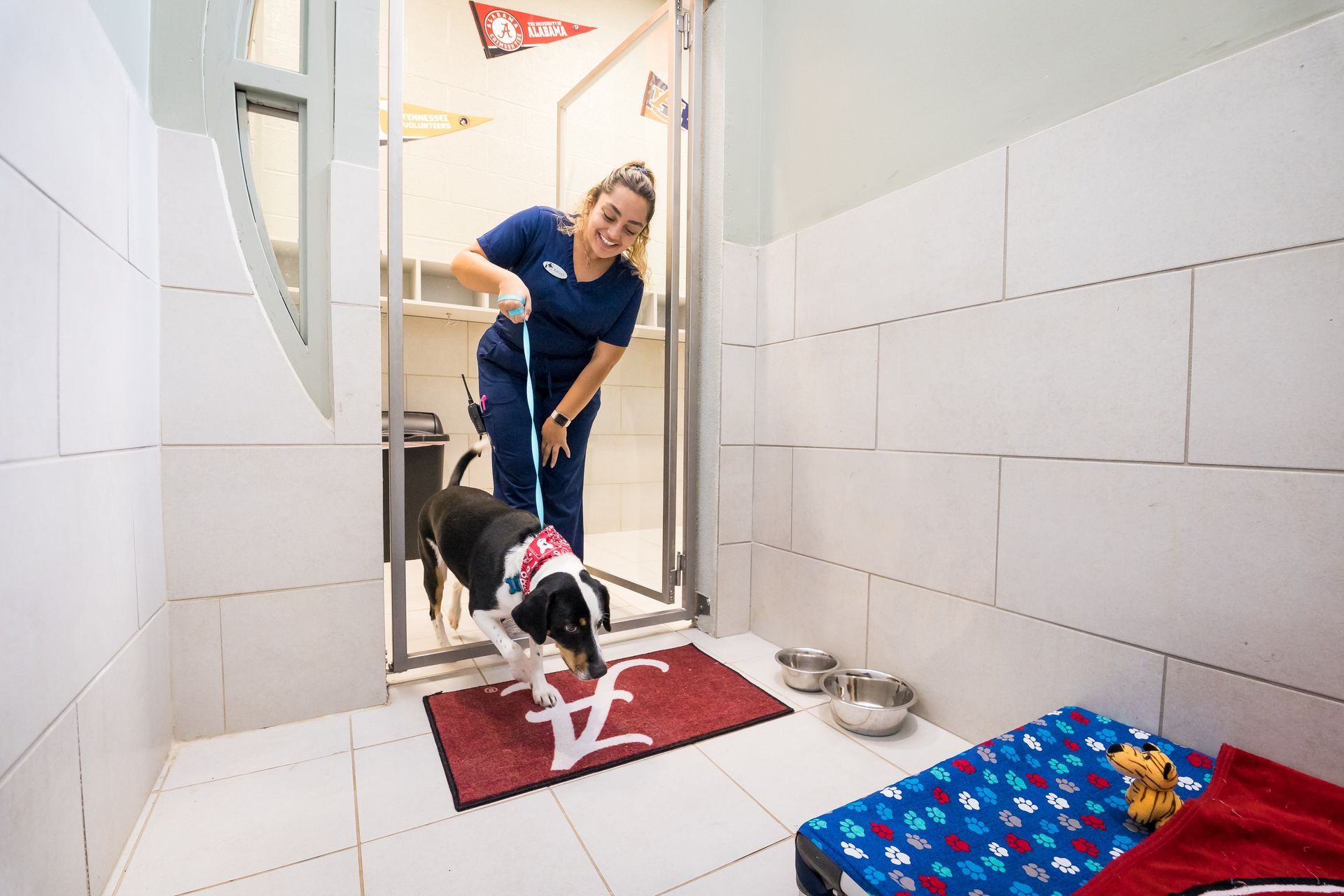Woman with a leash leading a dog from a dog kennel; the kennel has a bed, bowls, and a welcome mat.