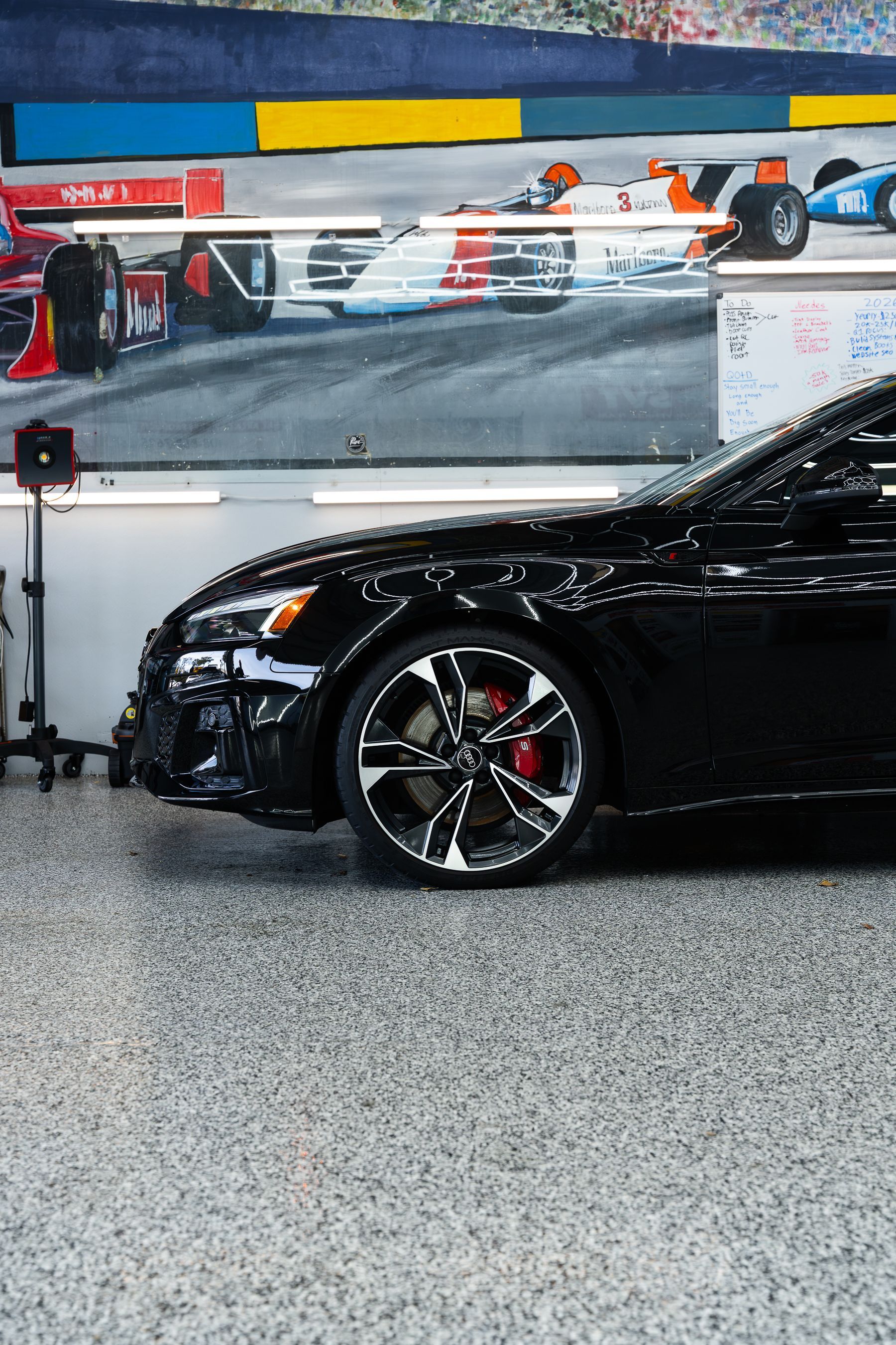 A shiny black car with red brake calipers parked on a speckled floor in front of a wall mural of racing cars.