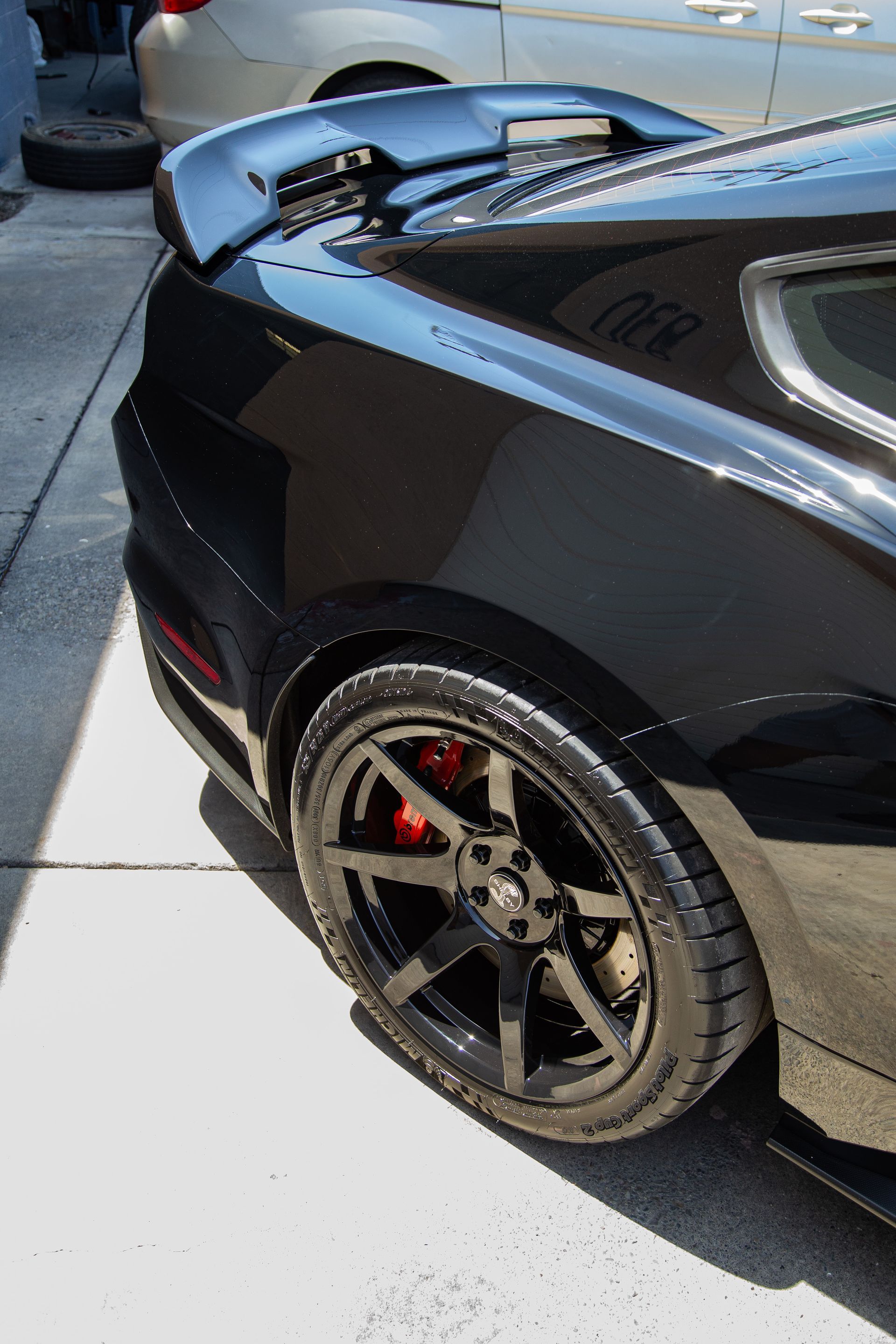 A black Ford Mustang with a rear spoiler, black wheels, and visible red brake calipers parked on pavement.