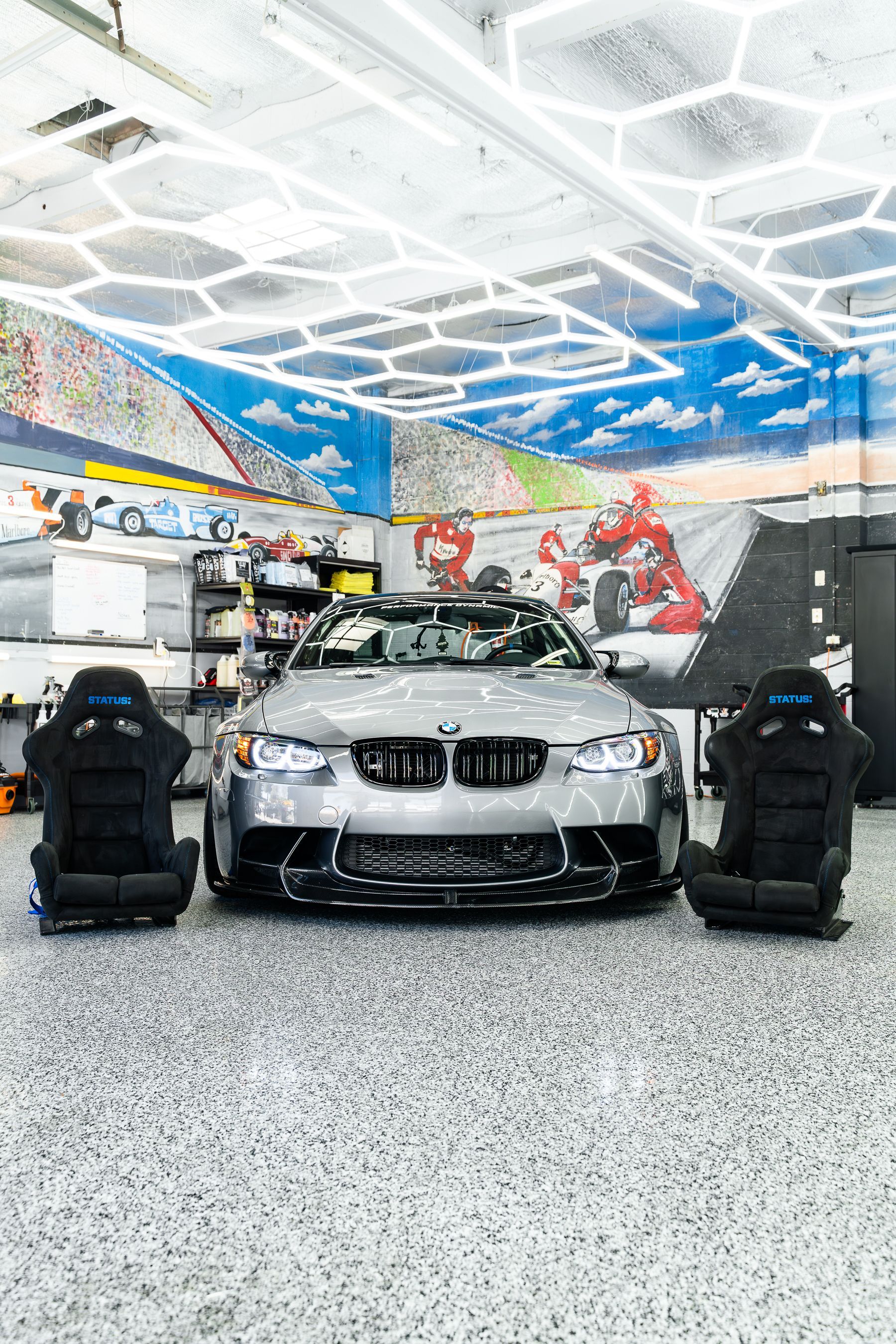 A silver BMW sports car parked in a garage with a honeycomb ceiling, wall mural, and two black racing seats in front.