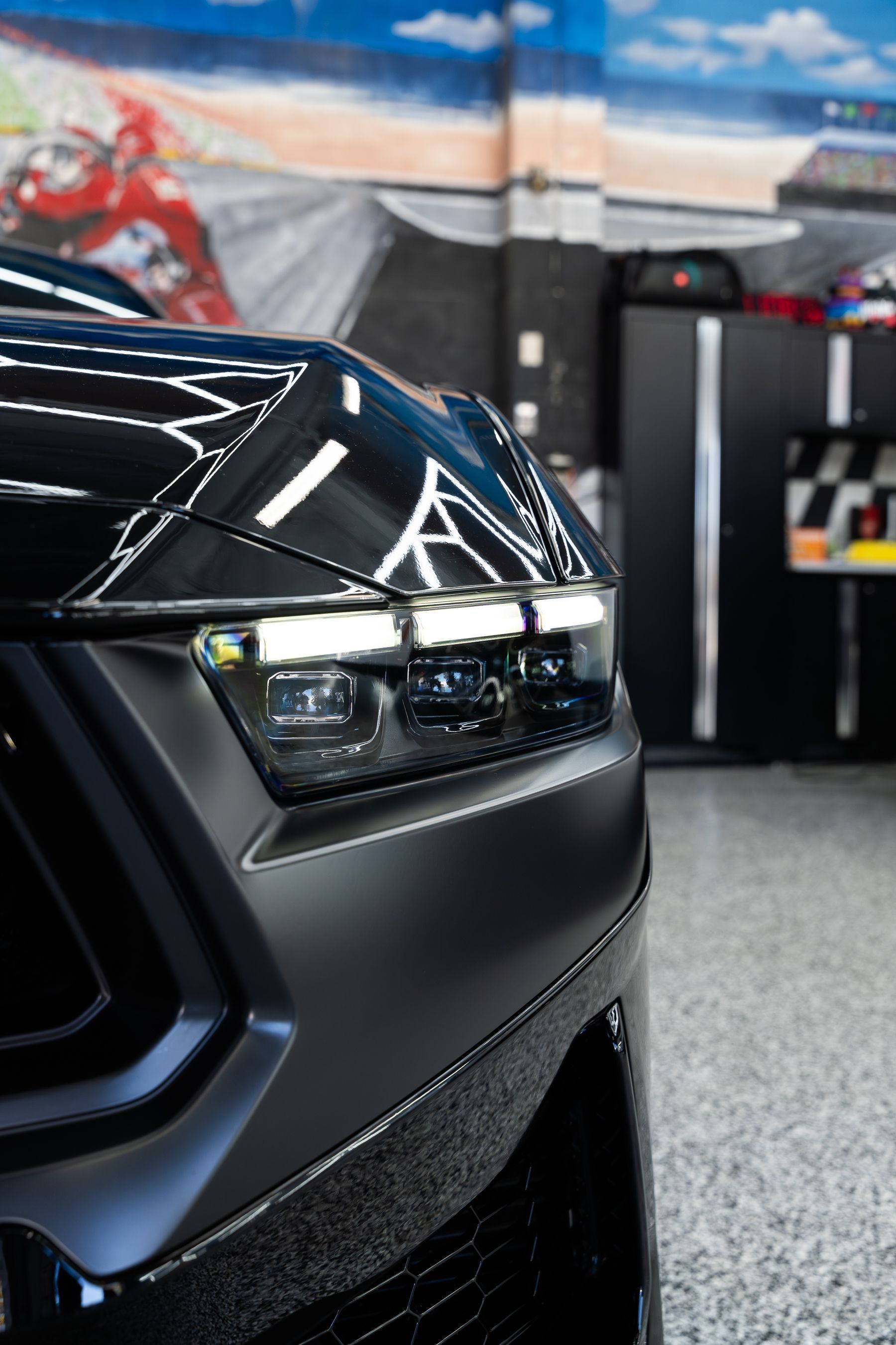 A close-up of a black car's modern LED headlight and matte hood inside a garage with a mural in the background.